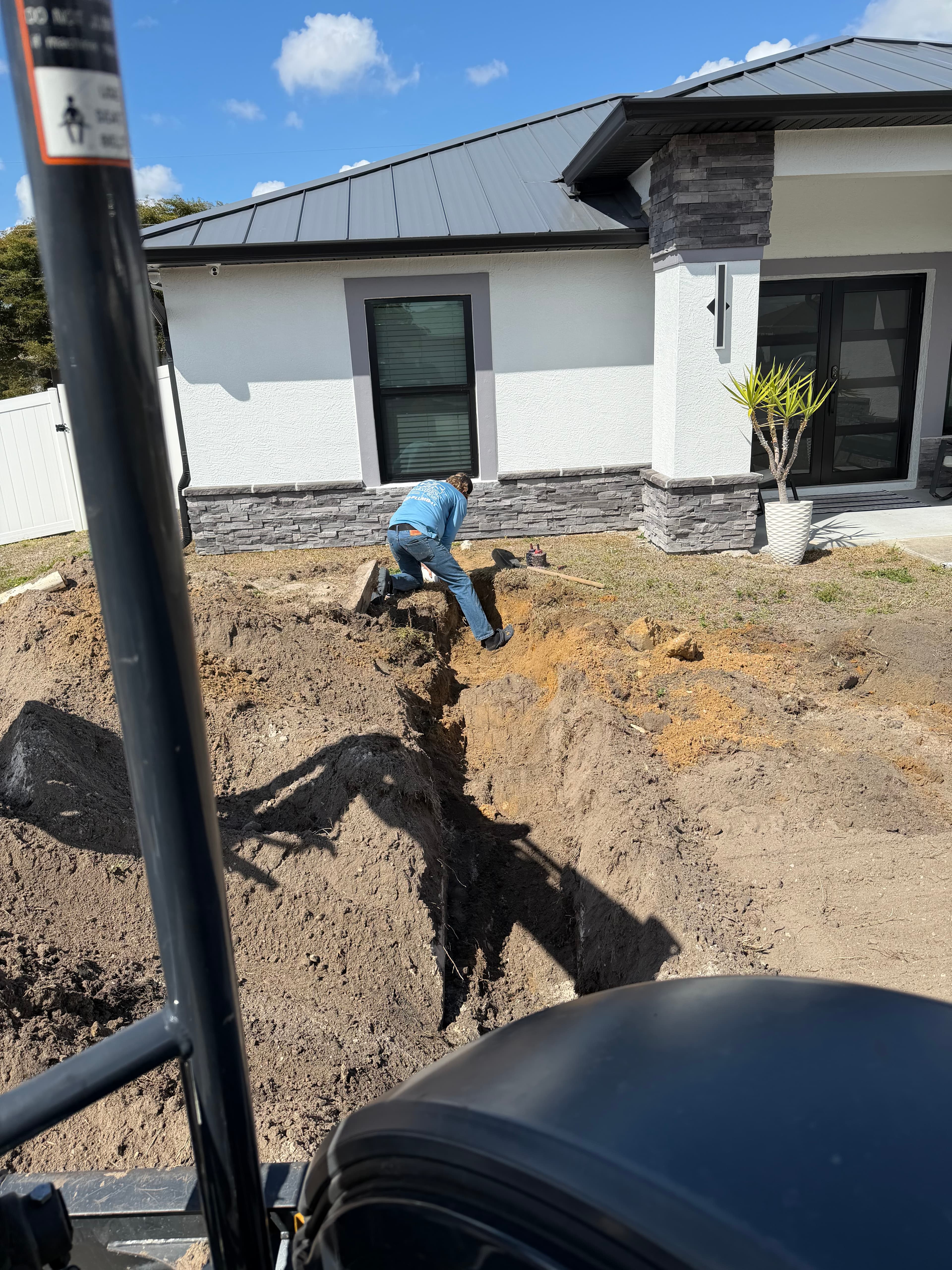 Excavation along residential property for sewer line installation — crew member in trench